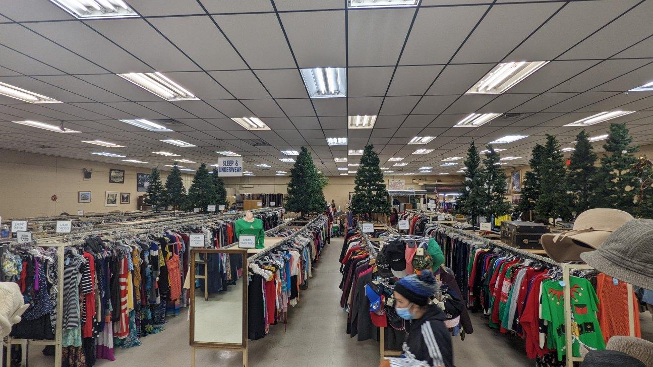 Clothing racks in a thrift store, Christmas trees in the center, and a person in a mask.