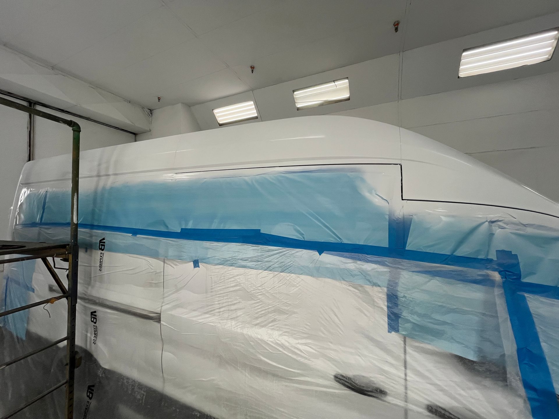 Un gros véhicule blanc recouvert de plastique de protection et de ruban adhésif bleu de peintre à l'intérieur d'une cabine de peinture industrielle.