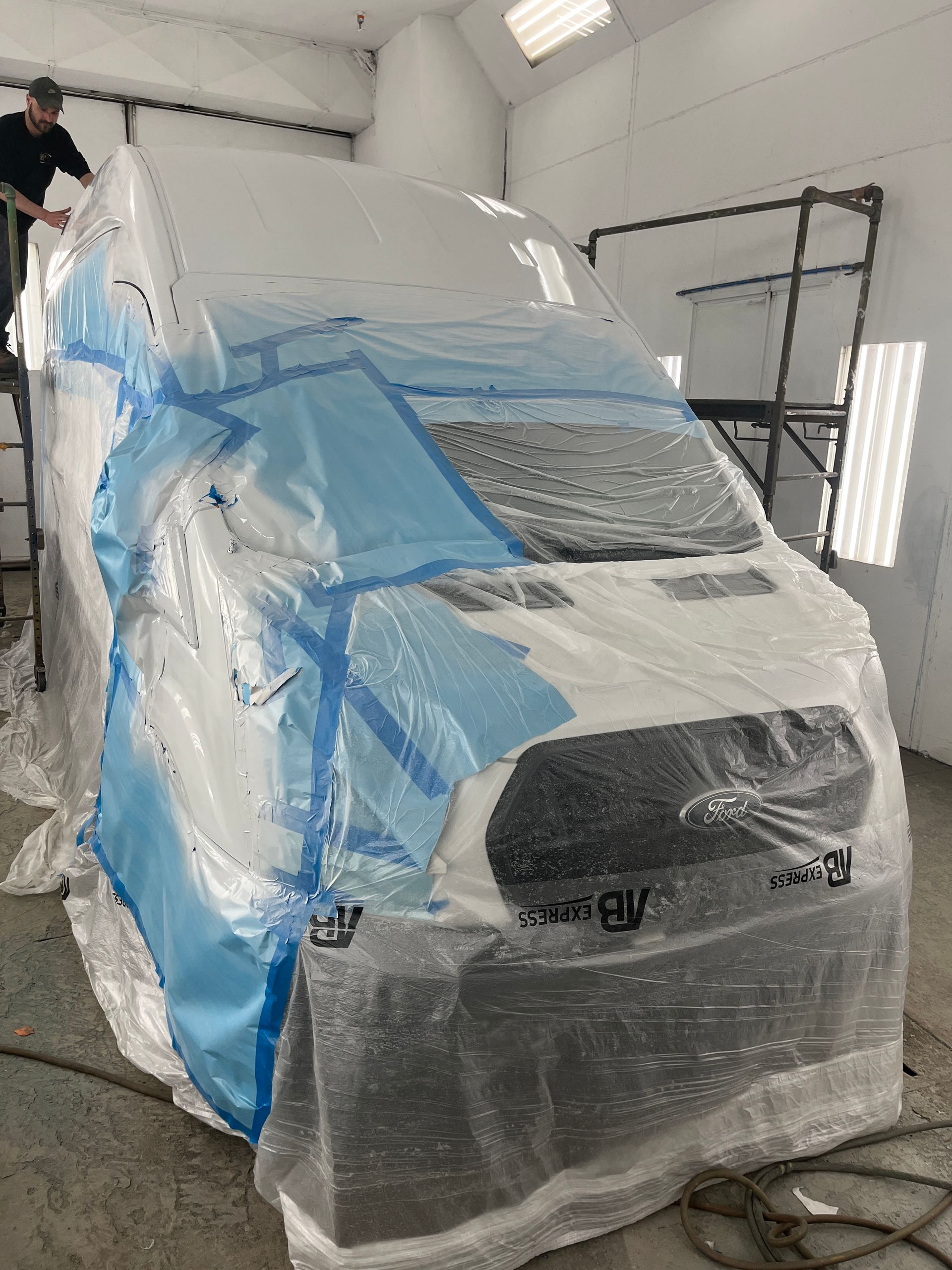 Une camionnette recouverte de bâches en plastique et de ruban adhésif bleu dans un atelier, une personne qui pose le ruban adhésif.