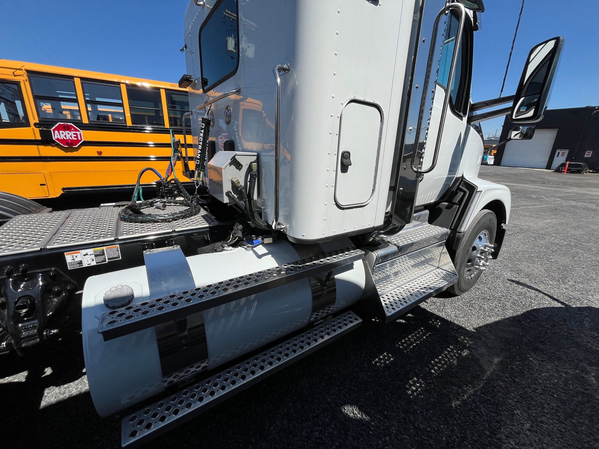 Un semi-remorque blanc est garé à l'extérieur, à côté d'un autobus scolaire jaune. Journée ensoleillée.