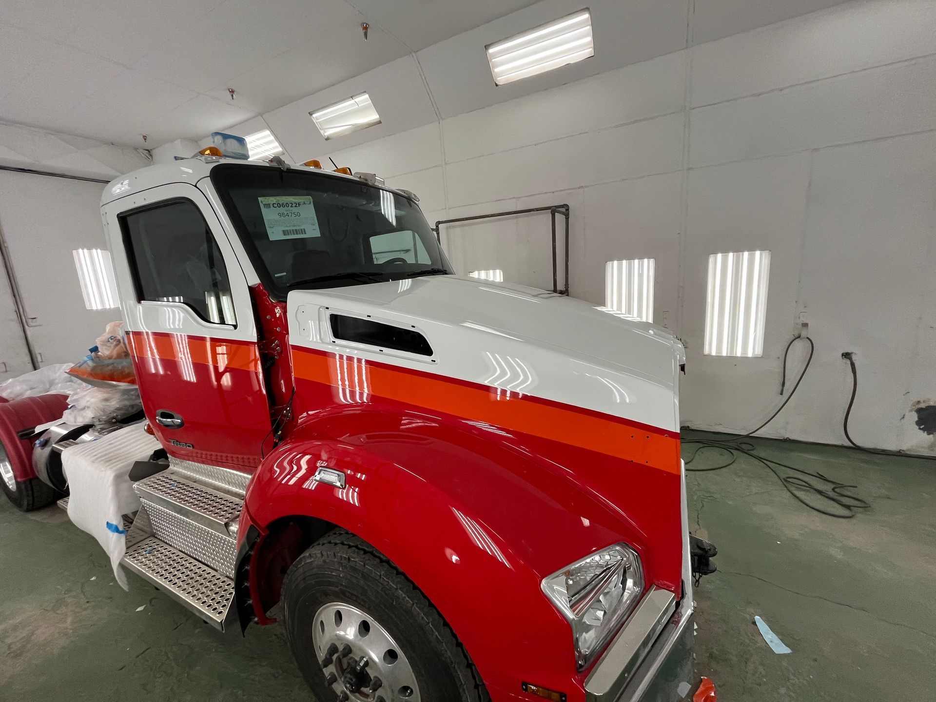 Un semi-remorque rouge, blanc et orange dans une cabine de peinture baignée de lumière.
