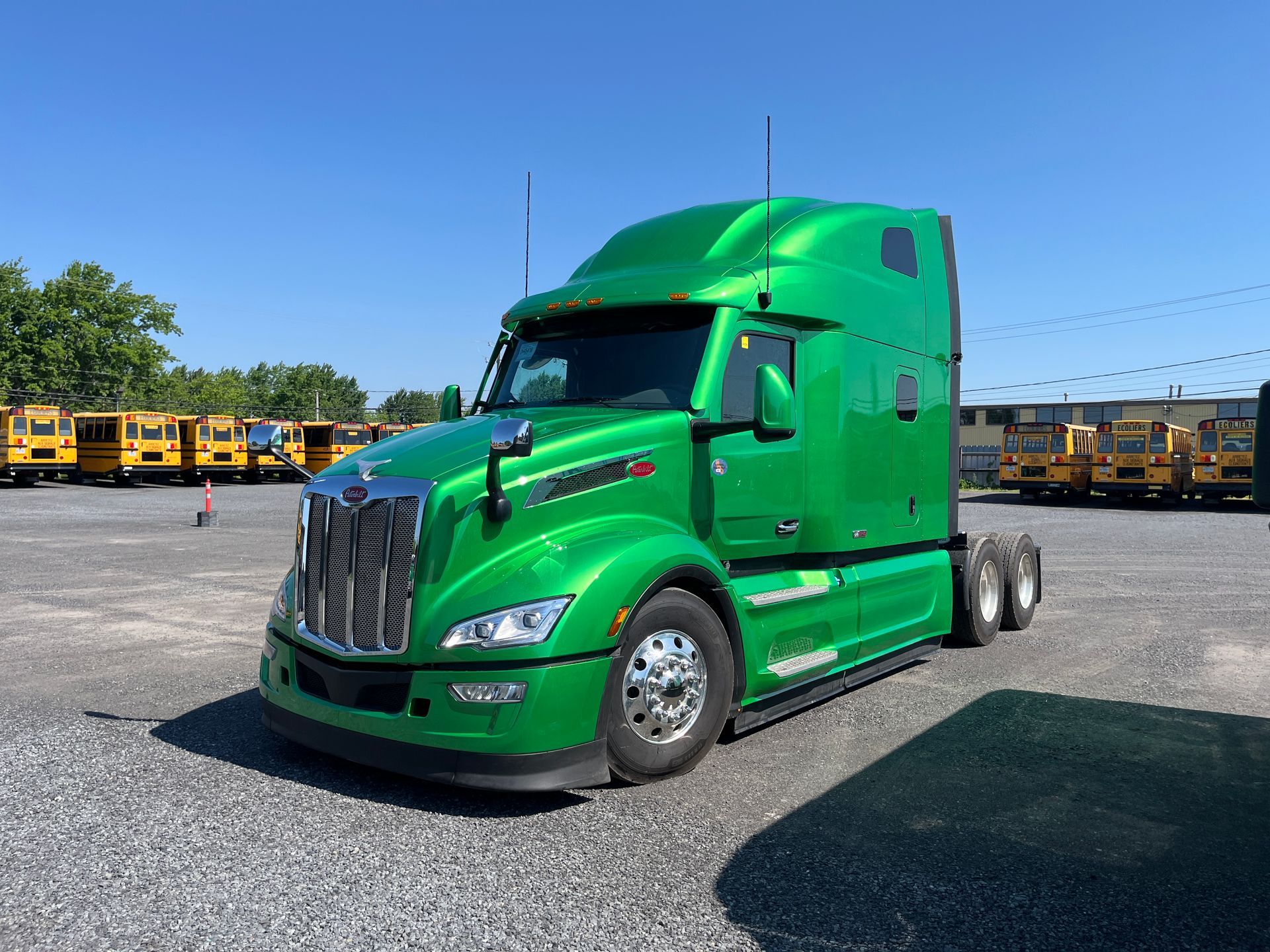 Semi-remorque vert garé sur un terrain en gravier, avec des autobus scolaires en arrière-plan. Journée ensoleillée.