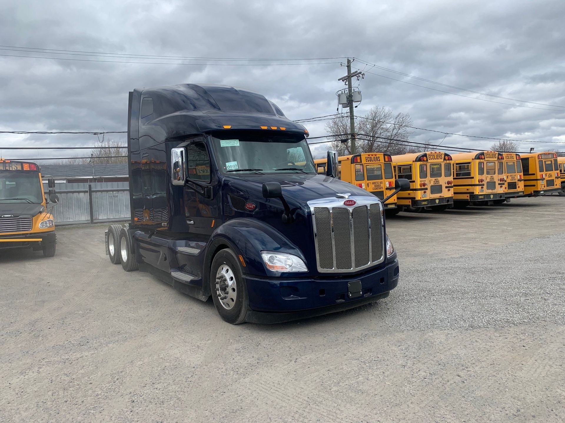 Semi-camion bleu foncé stationné sur un terrain en gravier, avec des autobus scolaires en arrière-plan par une journée nuageuse.
