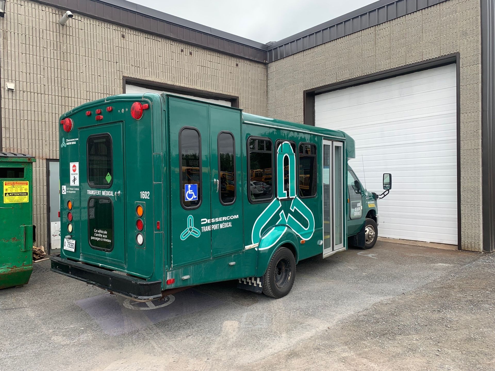 Un autobus vert adapté aux personnes à mobilité réduite est stationné devant un bâtiment doté de grandes portes sectionnelles.