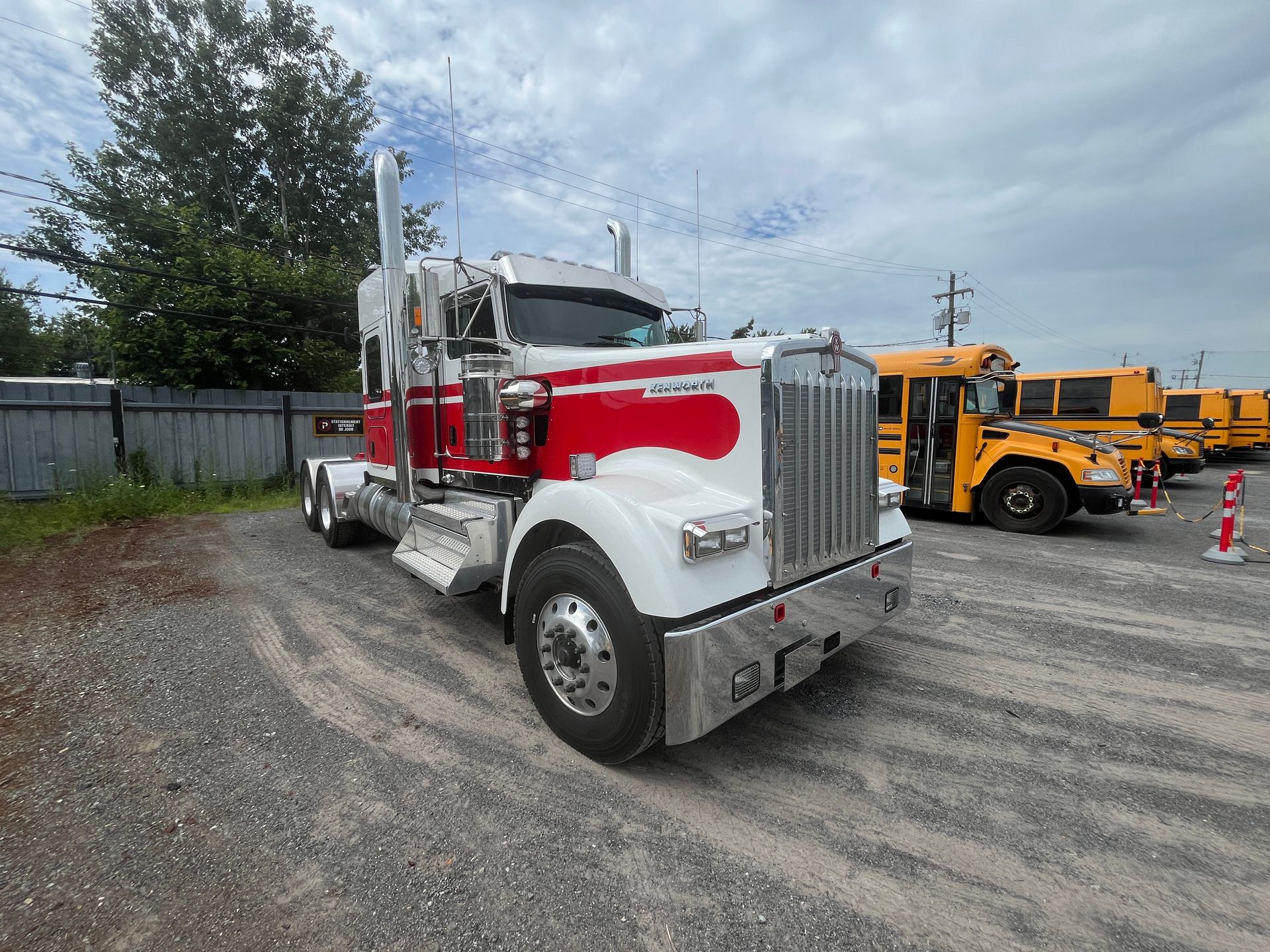 Un semi-remorque blanc et rouge est stationné sur un terrain en gravier, avec des autobus scolaires jaunes en arrière-plan, sous un ciel nuageux.
