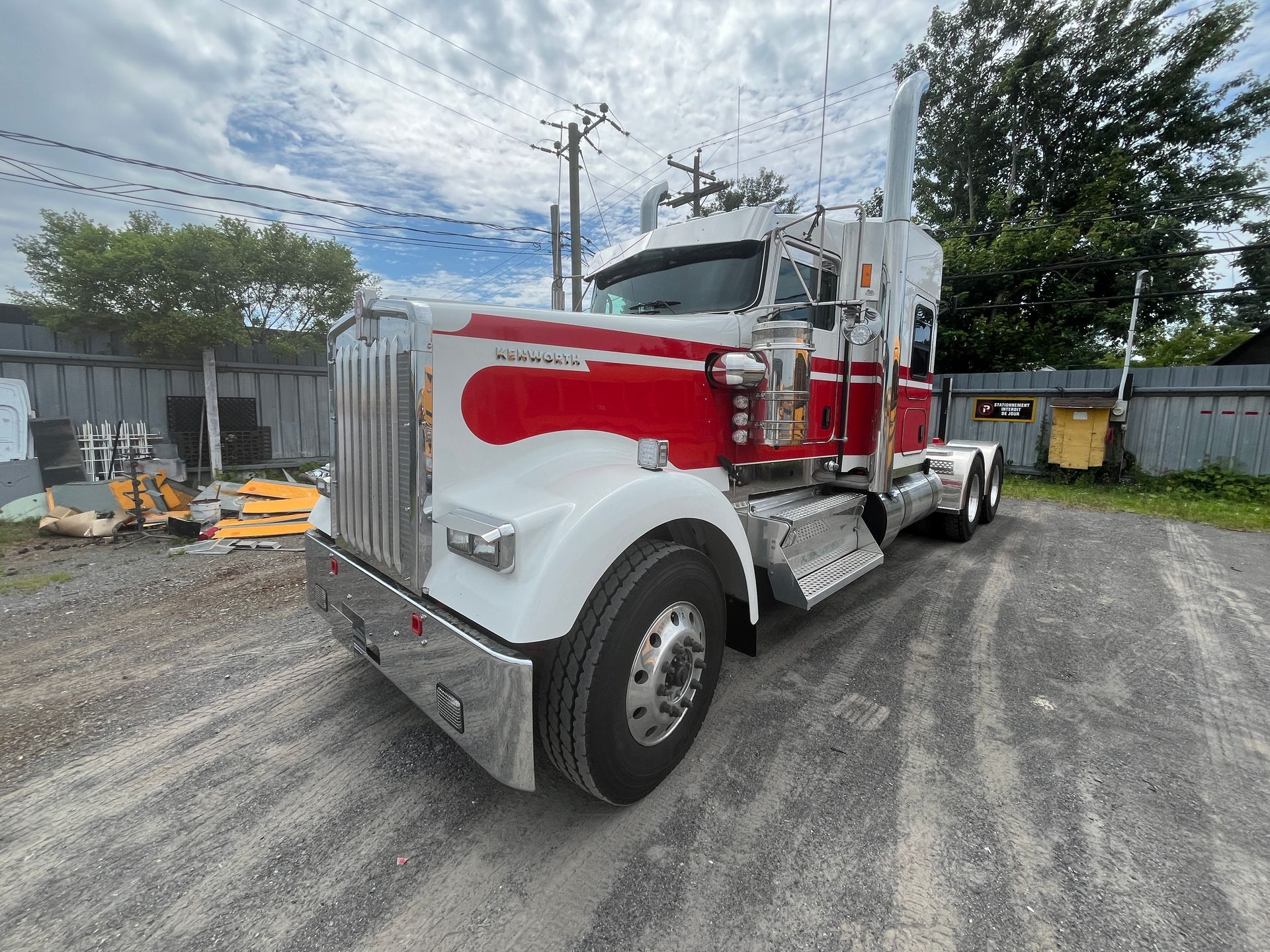 Semi-camion blanc et rouge garé sur du gravier, sur fond de ciel nuageux et de paysage industriel.