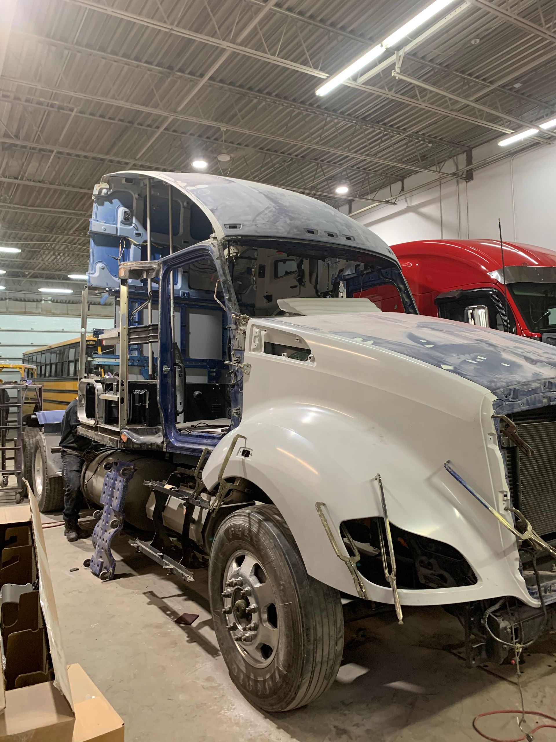 Châssis de semi-remorque en cours de réparation dans un atelier ; carrosserie blanche et bleue ; camion rouge visible en arrière-plan.