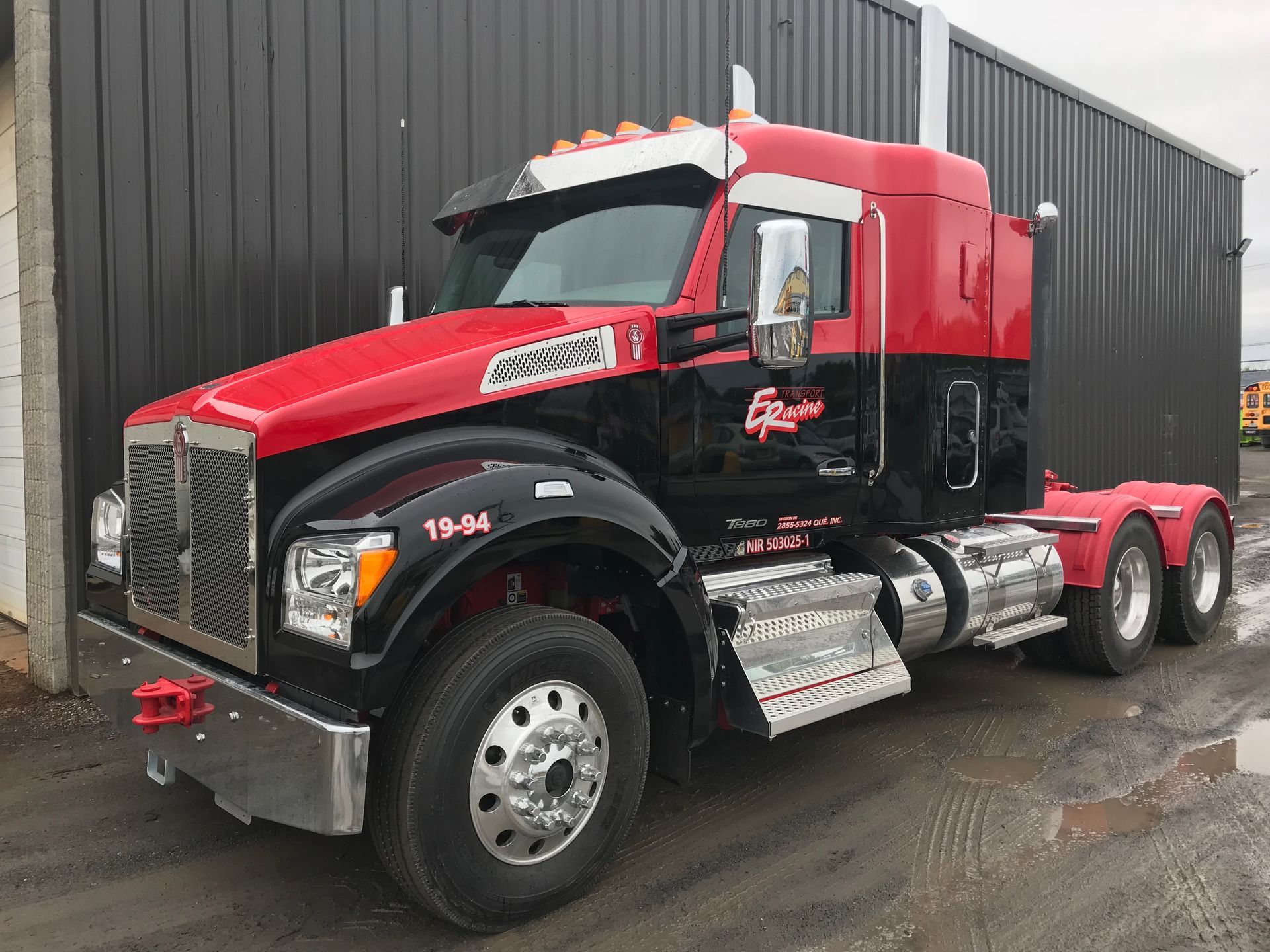 Un semi-remorque rouge et noir est garé devant un bâtiment gris.