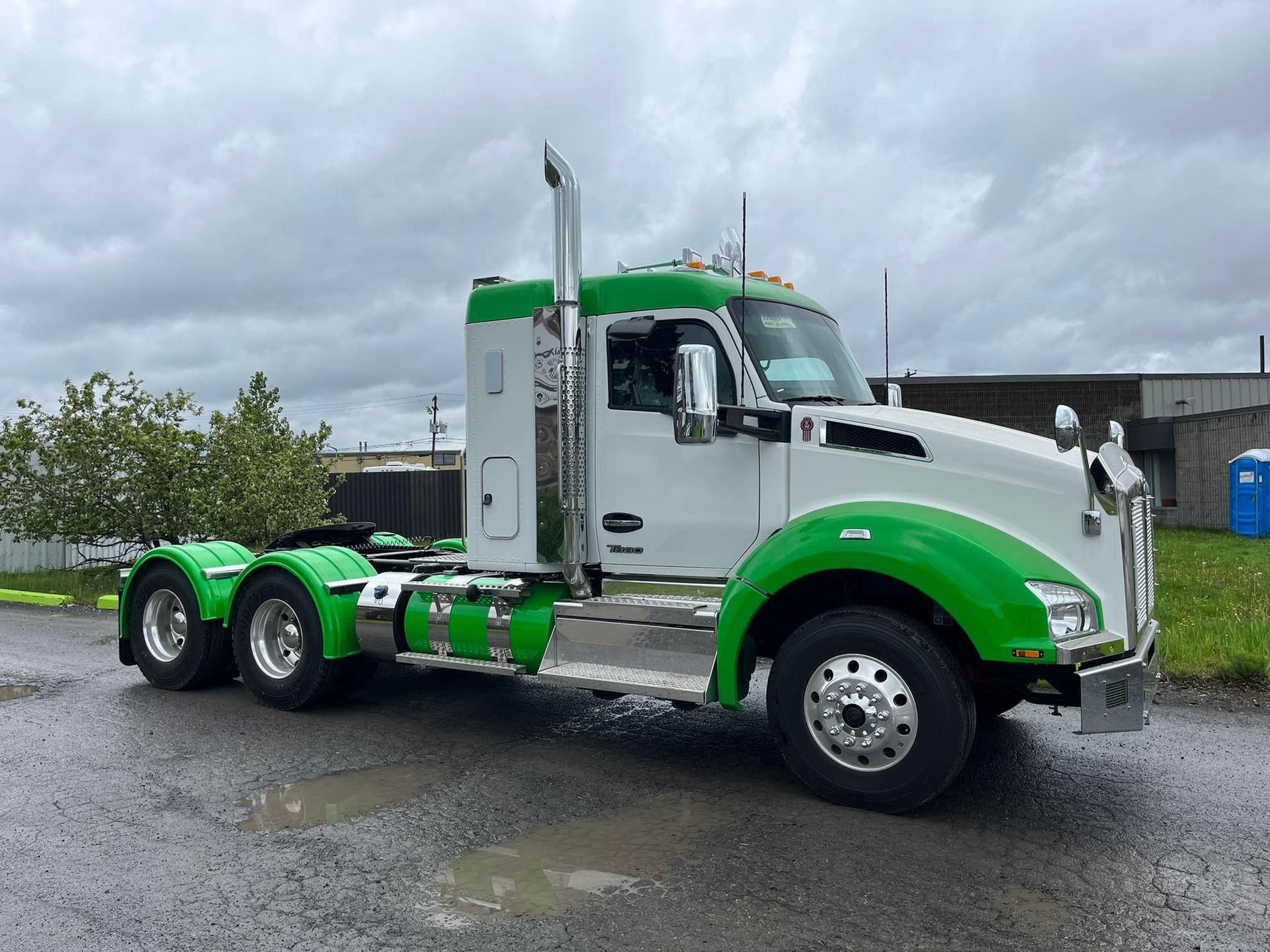 Semi-camion Kenworth blanc et vert stationné sur une chaussée mouillée avec un ciel couvert.