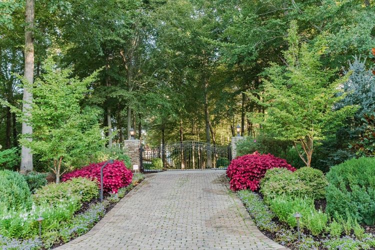 A brick driveway leading to a gate in a garden surrounded by trees and flowers.