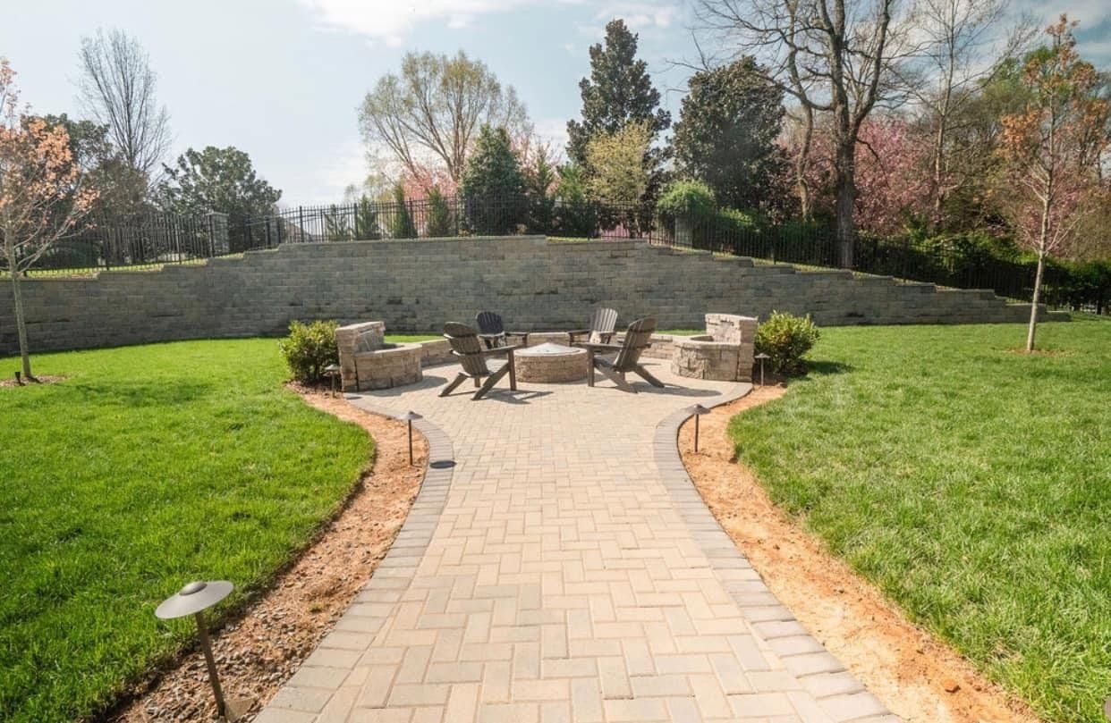 A brick walkway leading to a fire pit in a lush green yard.