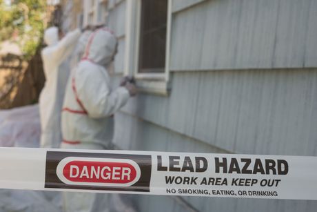 A man in a protective suit is standing in front of a lead hazard sign.