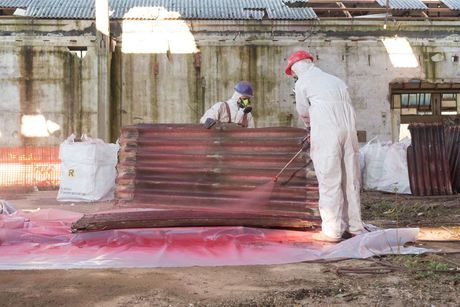 Two men in white overalls are spraying red paint on a pile of wood.