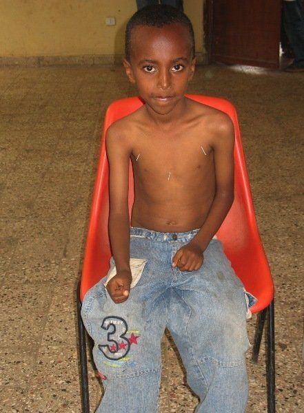 A shirtless young boy sits in a red chair with the number 3 on his pants