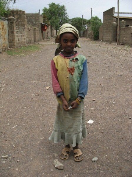 A little girl wearing a colorful shirt and skirt is standing on a dirt road