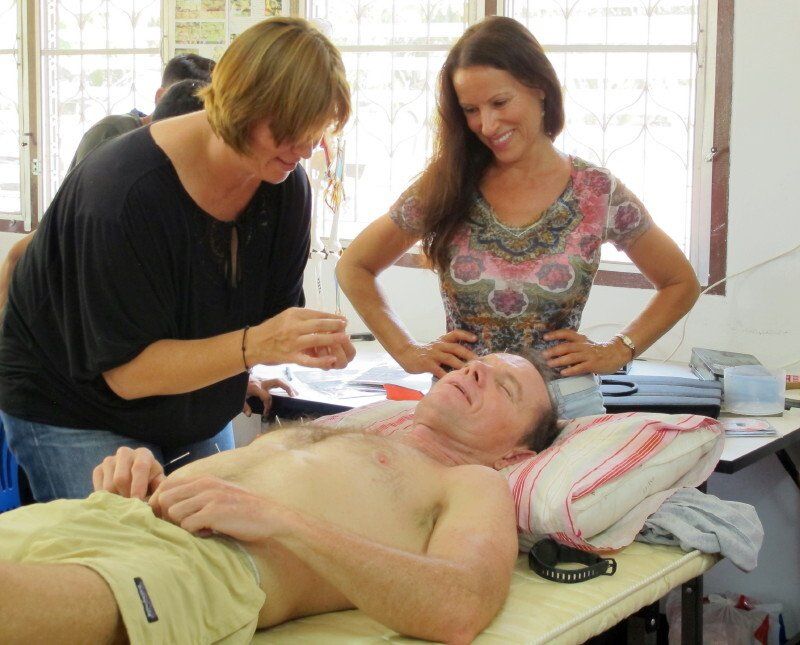A shirtless man is laying on a table with two women standing around him