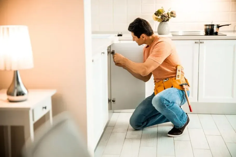 A person wearing a tool belt kneeling on a kitchen floor while repairing a white cabinet door.