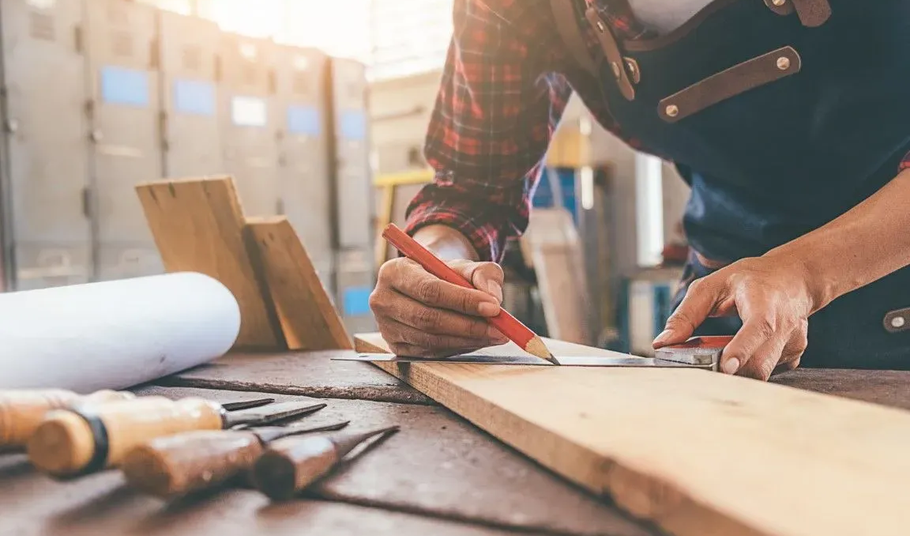 A close-up of a woodworker in an apron using a pencil and square to mark measurements on a wooden board in a workshop.
