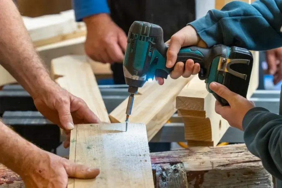 Hands hold a cordless drill, driving a screw into a wooden board on a workbench, with another person assisting nearby.