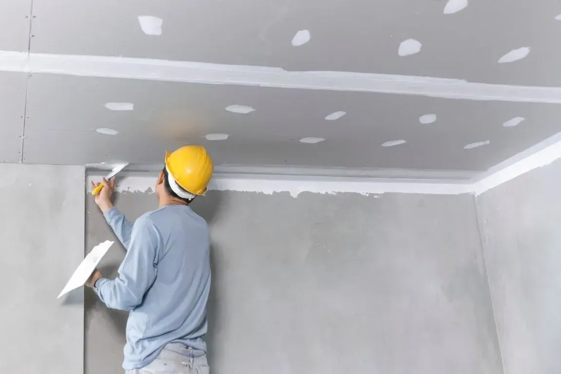 A worker in a yellow hard hat applies joint compound to a ceiling and wall corner with a putty knife and hawk.