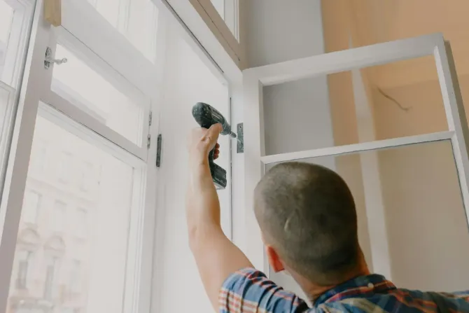 A person in a plaid shirt uses a power drill to install a white window frame indoors.