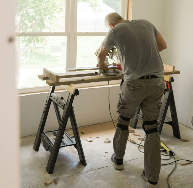 A person wearing knee pads stands at a makeshift wooden workbench, operating a miter saw in a room under construction.