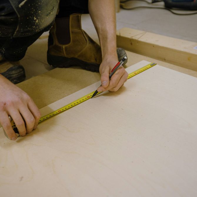 A person uses a pencil and tape measure to mark a measurement on a light-colored wooden board in a workshop.