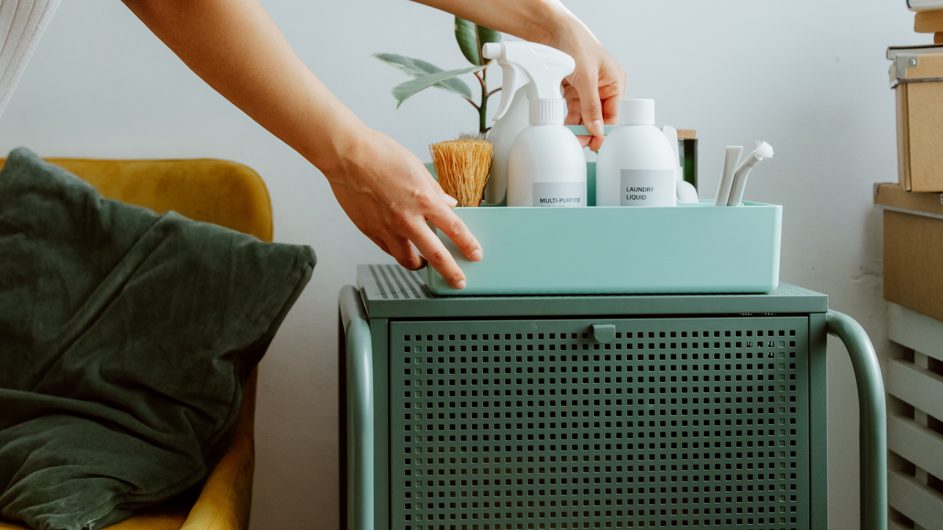 Person arranging cleaning supplies on a teal cart, next to a yellow chair and green blanket.