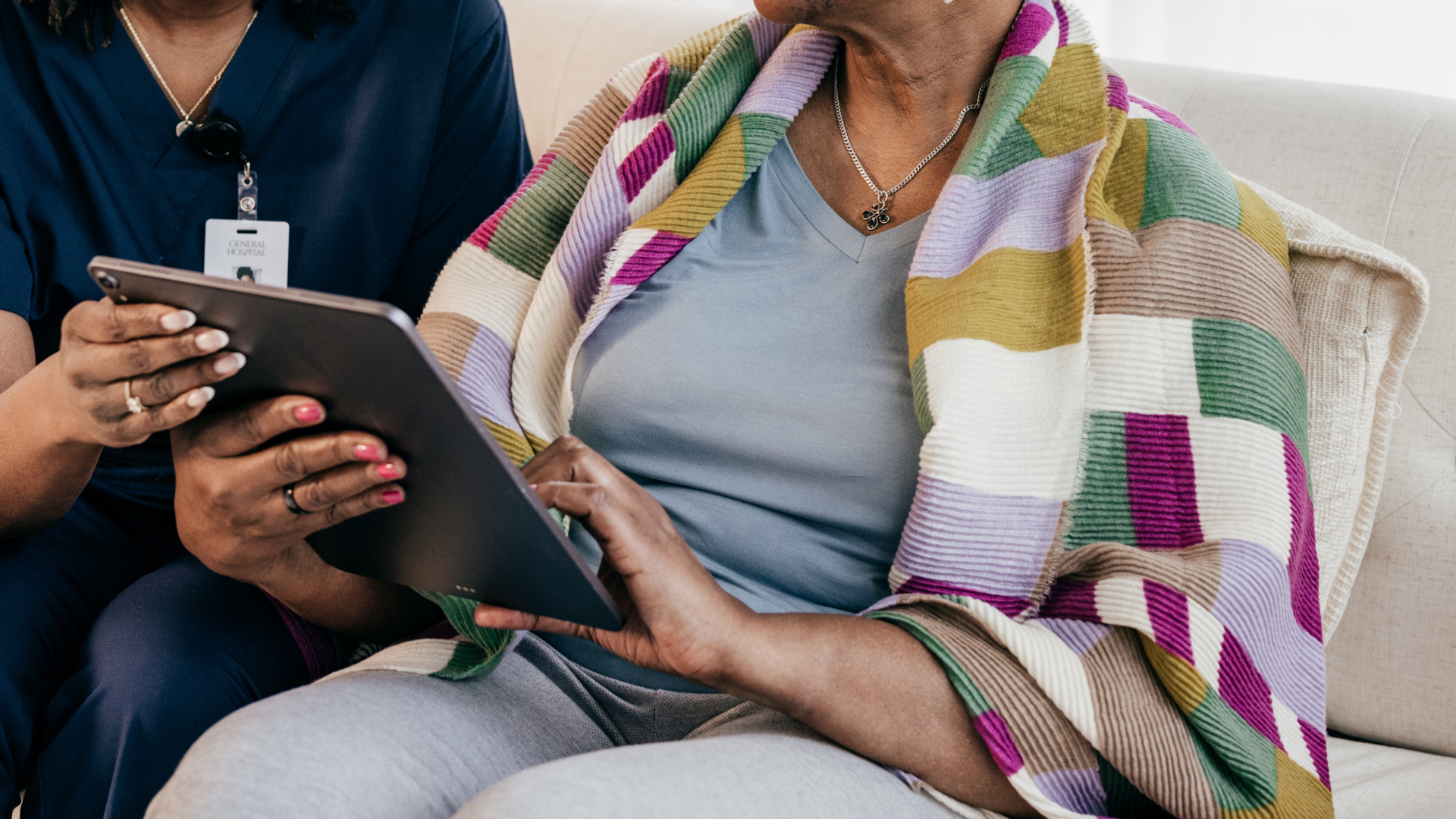 Nurse shows tablet to senior wrapped in a quilt on a couch.