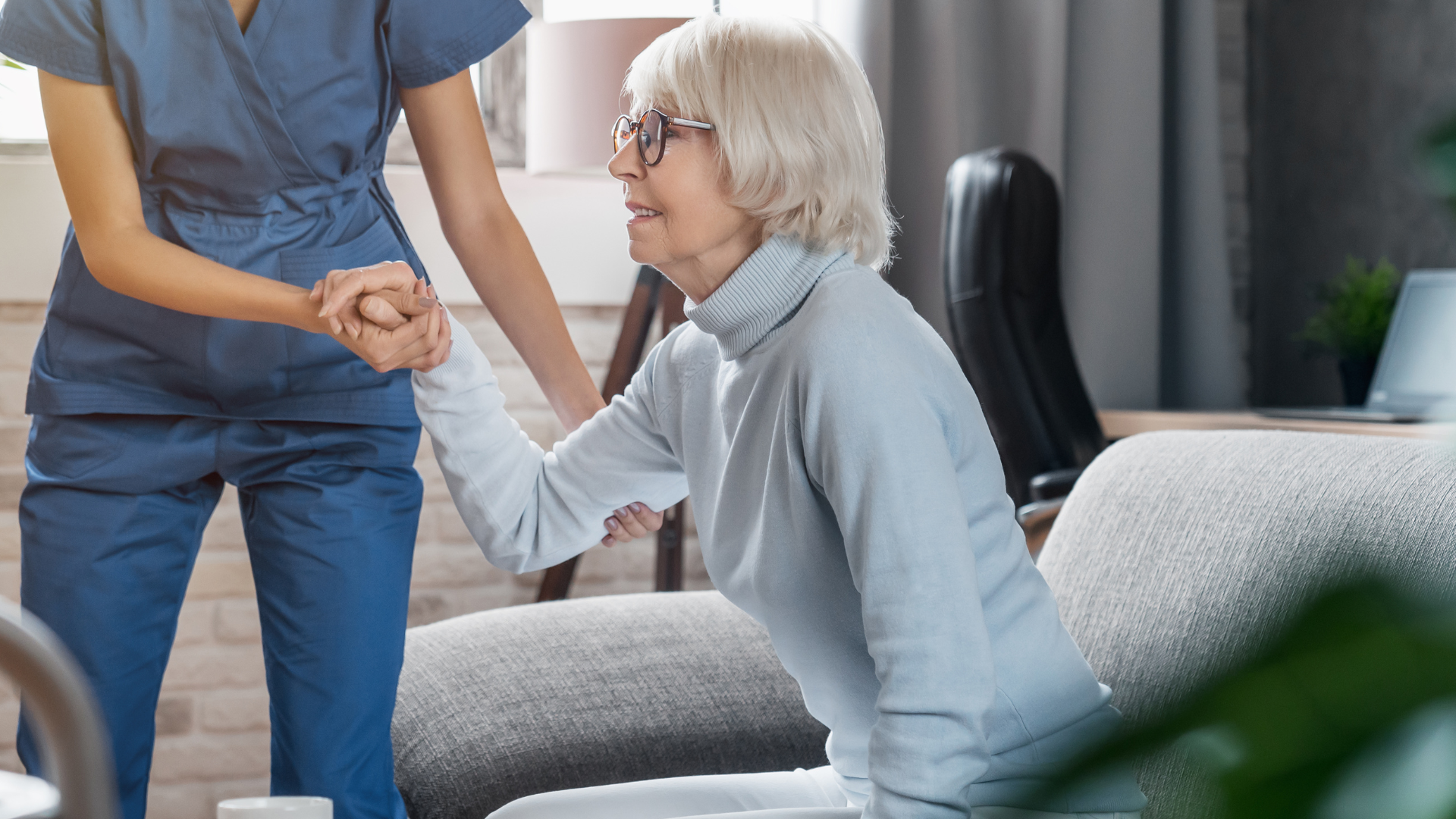 Nurse helping elderly woman rise from a couch. Woman in glasses smiles. Indoor setting.