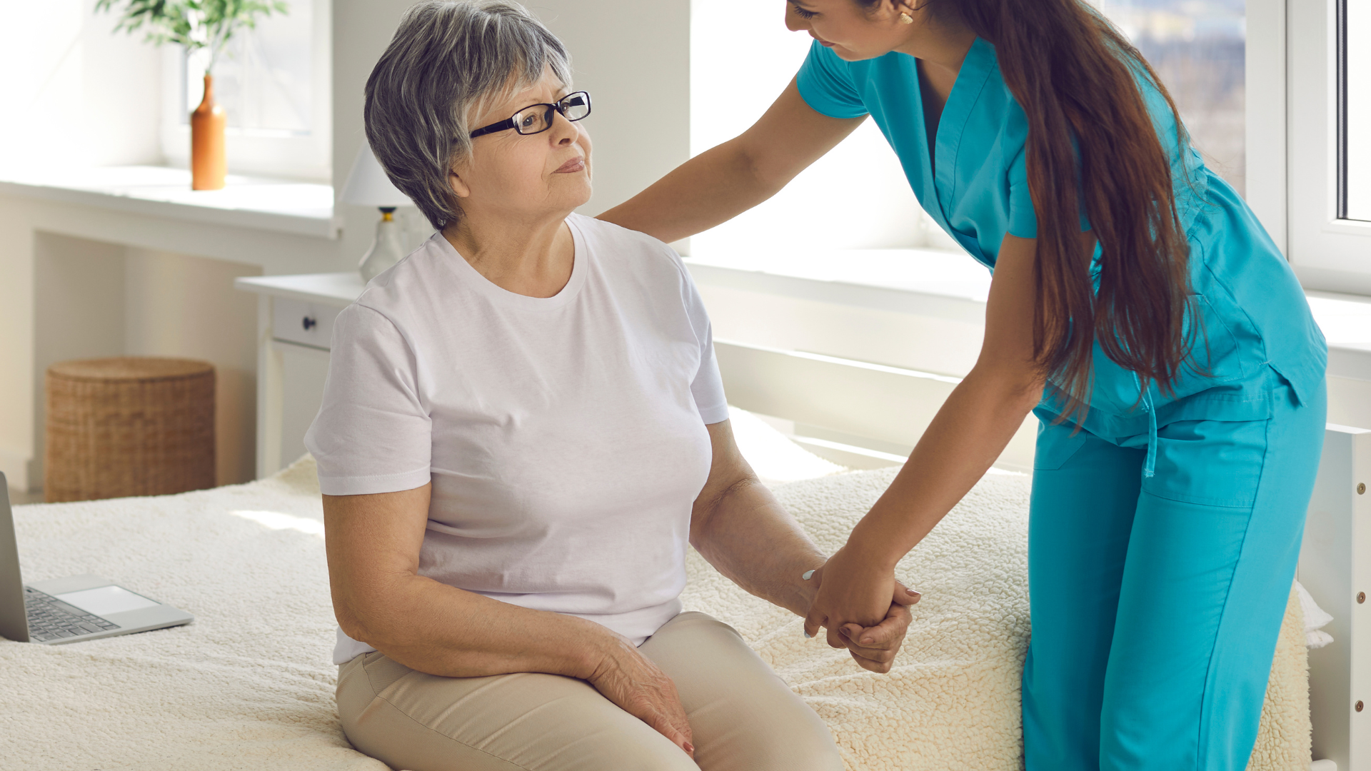 Caregiver in teal scrubs helps older woman in white shirt, seated on a bed.