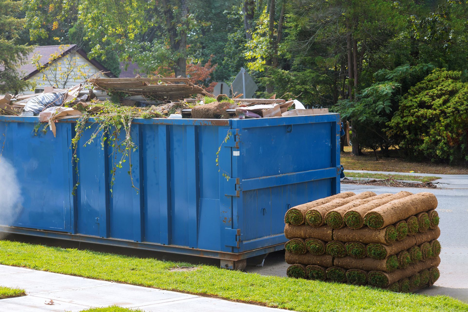 Blue dumpster filled with yard debris beside stacked rolls of sod.