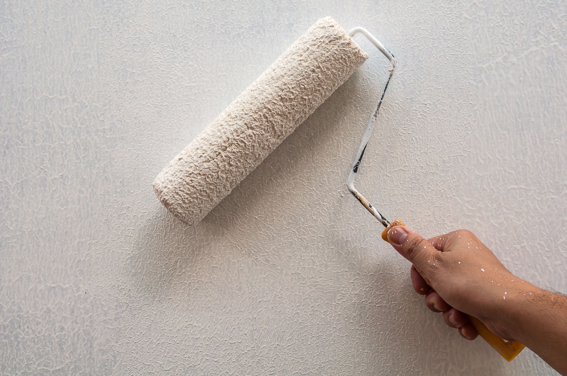 Hand using a paint roller to apply textured white paint to a wall.