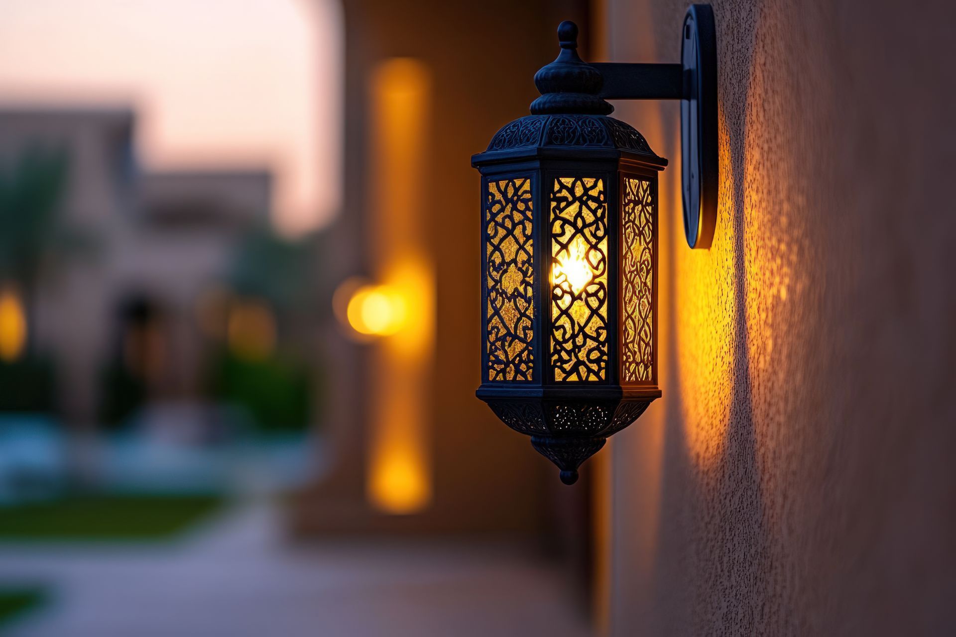 Ornate black lantern with warm light on a beige wall, blurred background of building at dusk.
