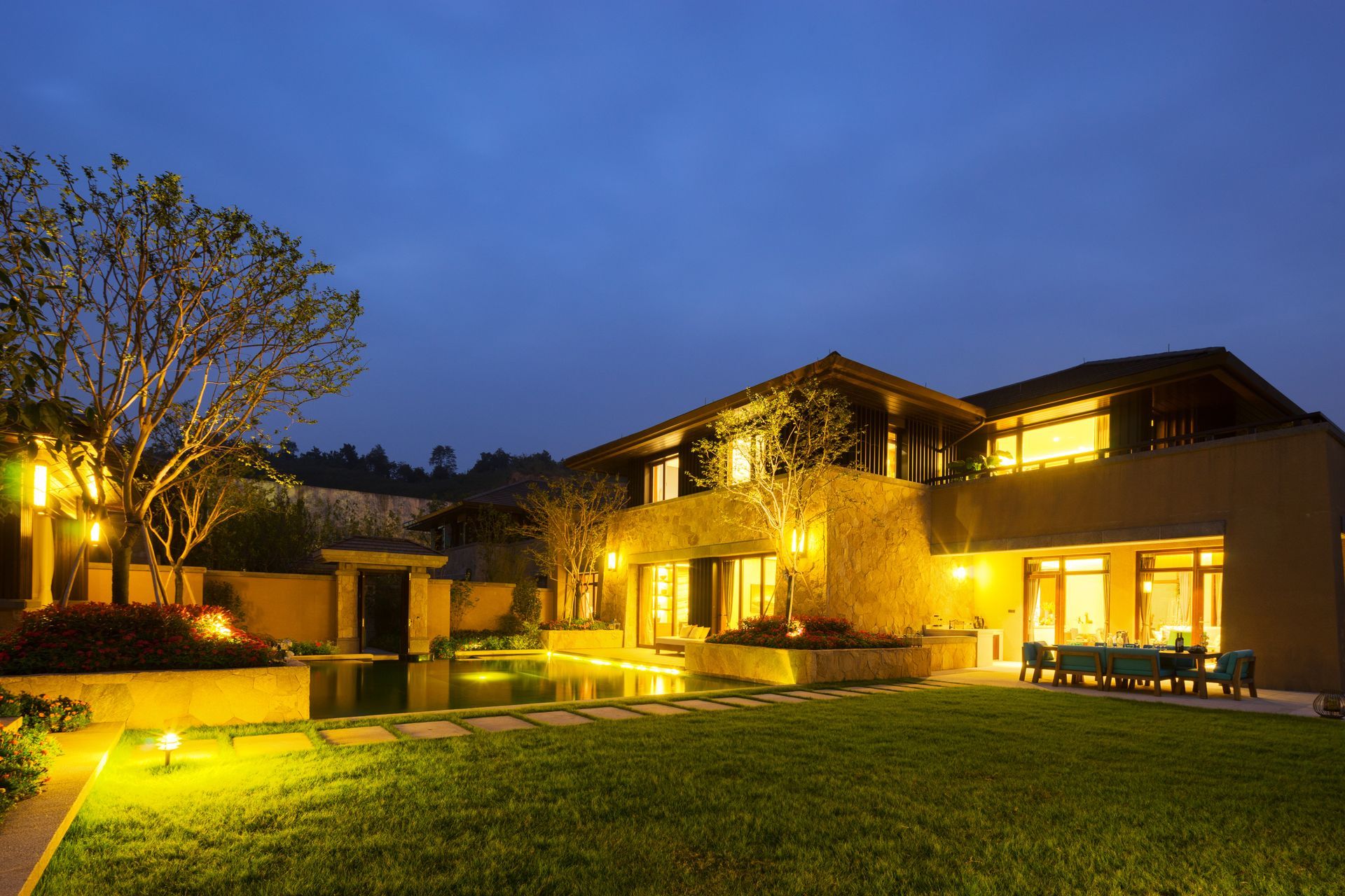Night view of a modern house with warm exterior lighting, a grassy yard, and a small pool.