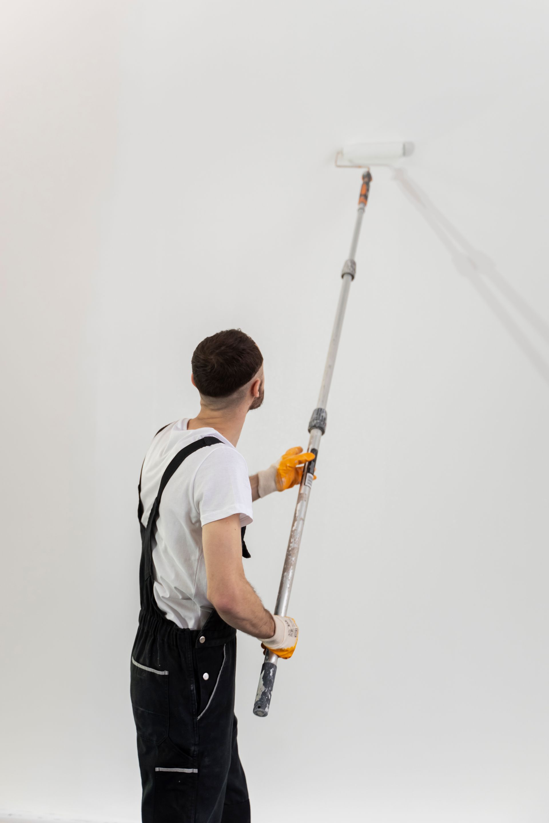 Person painting a white wall with a roller and extendable pole, wearing overalls and gloves.