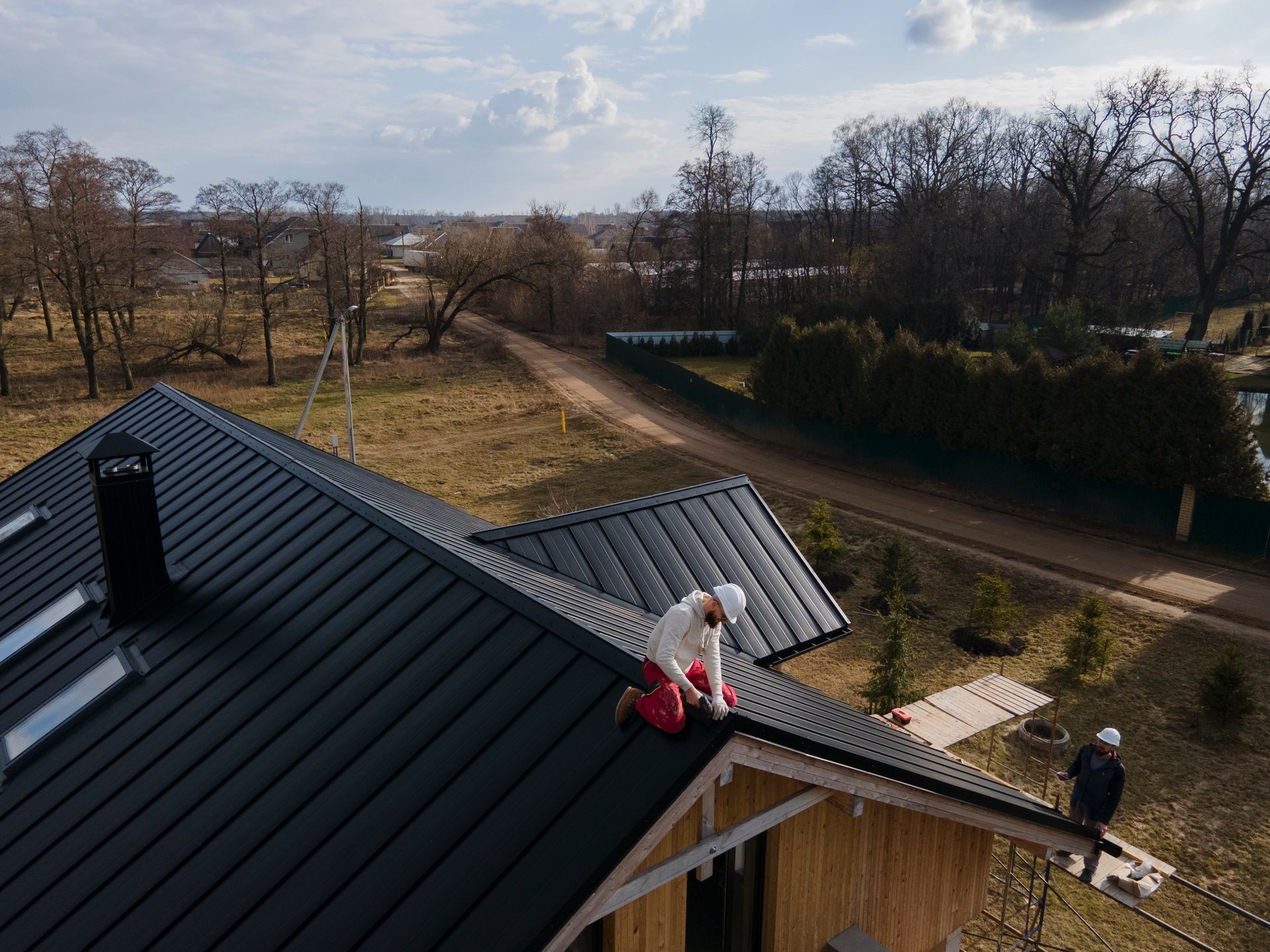 Two workers installing a dark metal roof on a house in a rural setting.