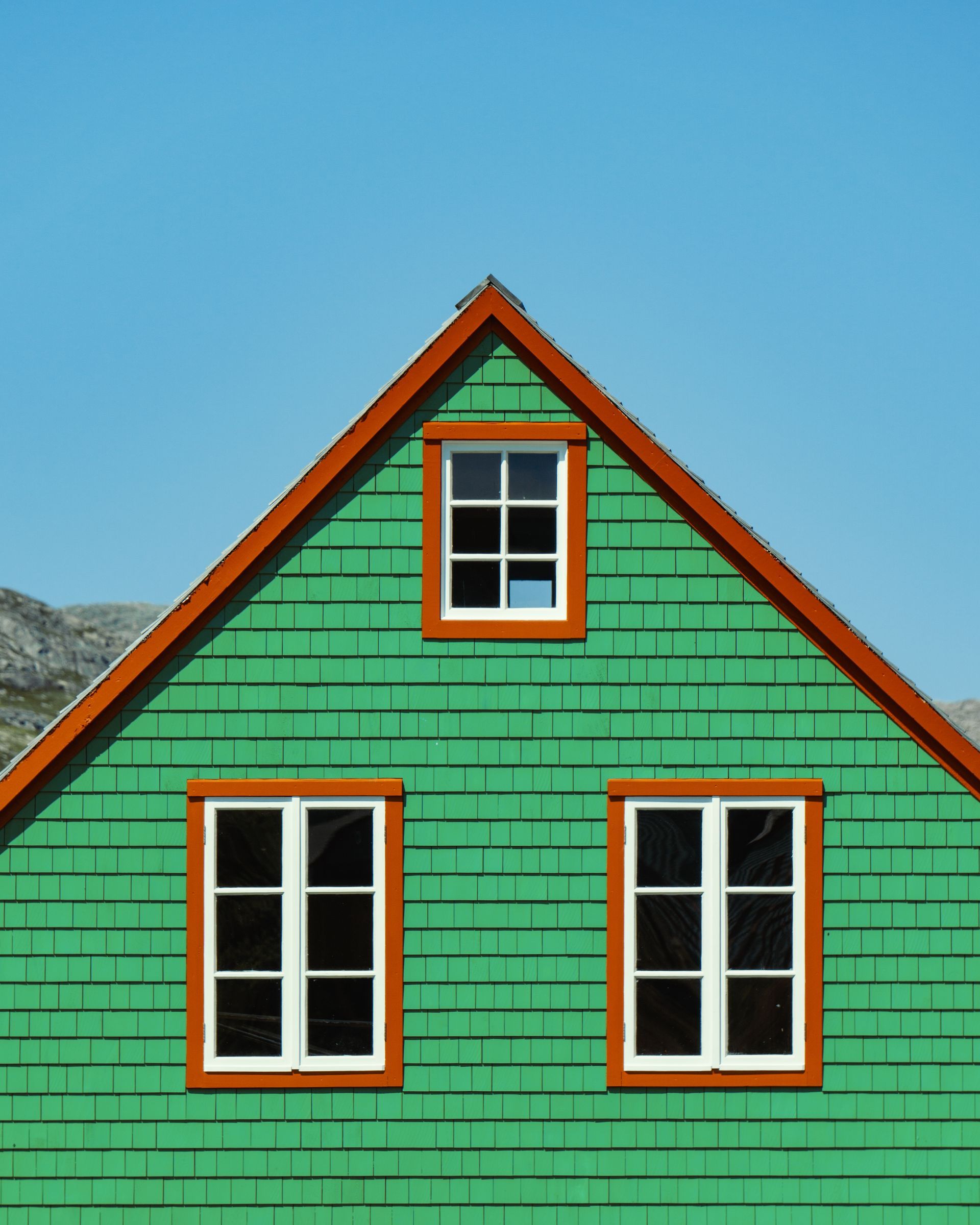 Green shingled house with orange trim and three white-framed windows under a blue sky.