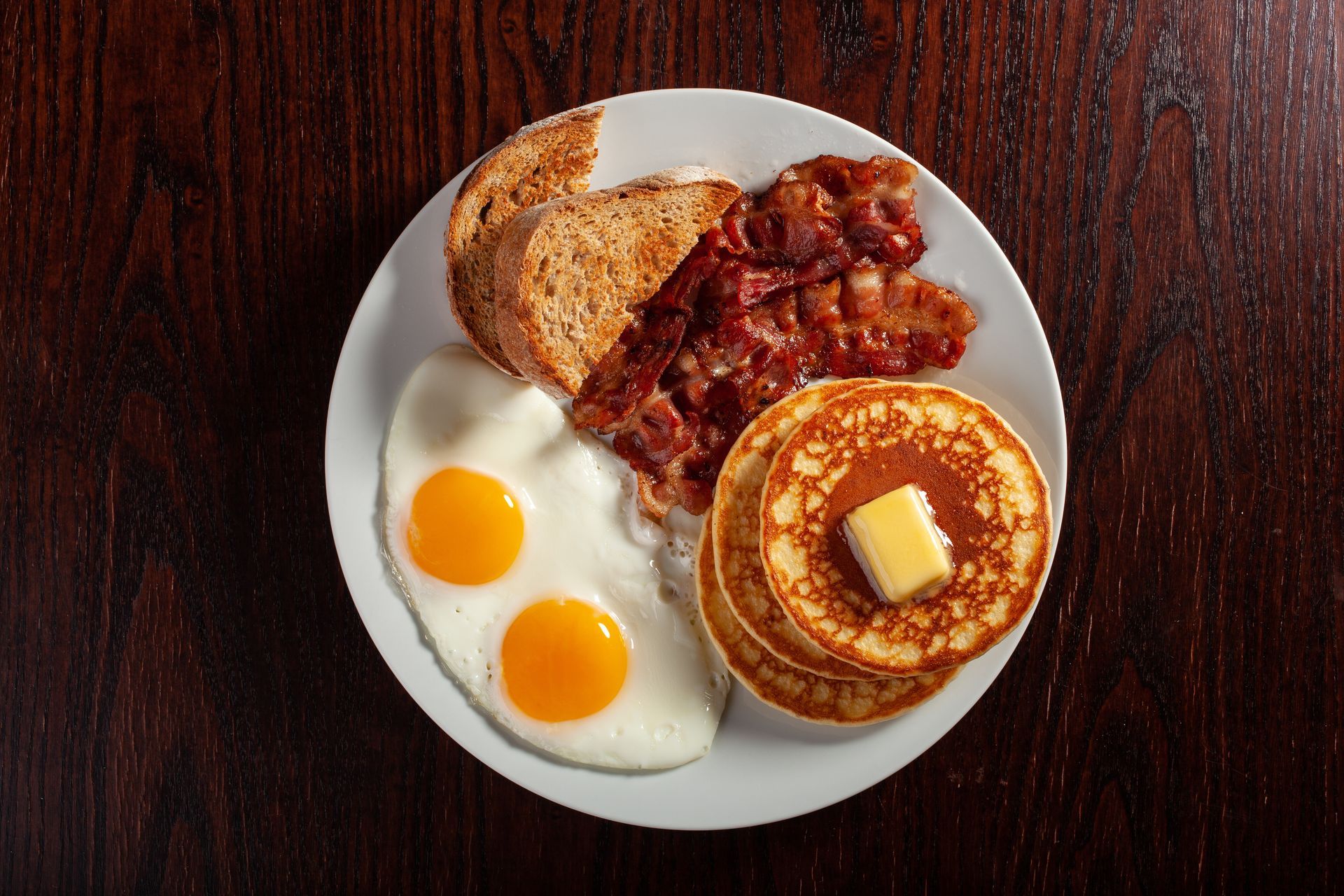 A plate of food with eggs , pancakes , bacon and toast on a wooden table.