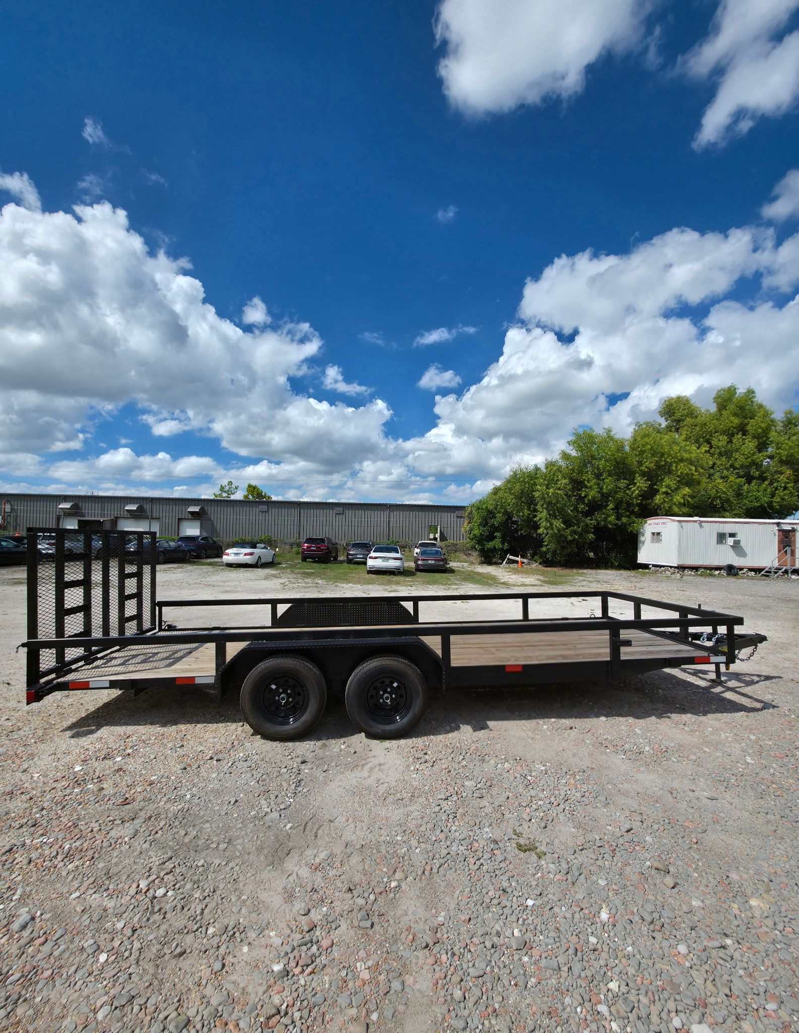 Black open-bed trailer on gravel, against blue sky with clouds, near building and trees.