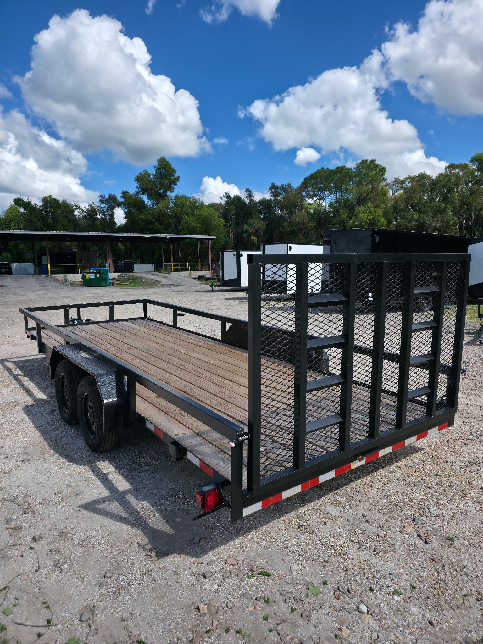 Black utility trailer with mesh sides on gravel, against a bright blue sky with clouds.