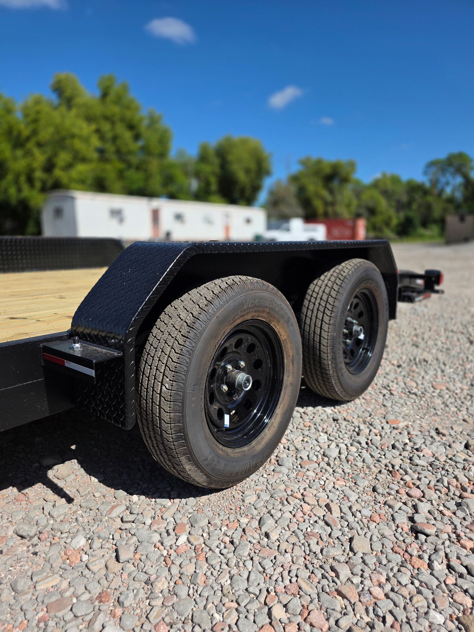 Black trailer with two tires on gravel with a blue sky background.