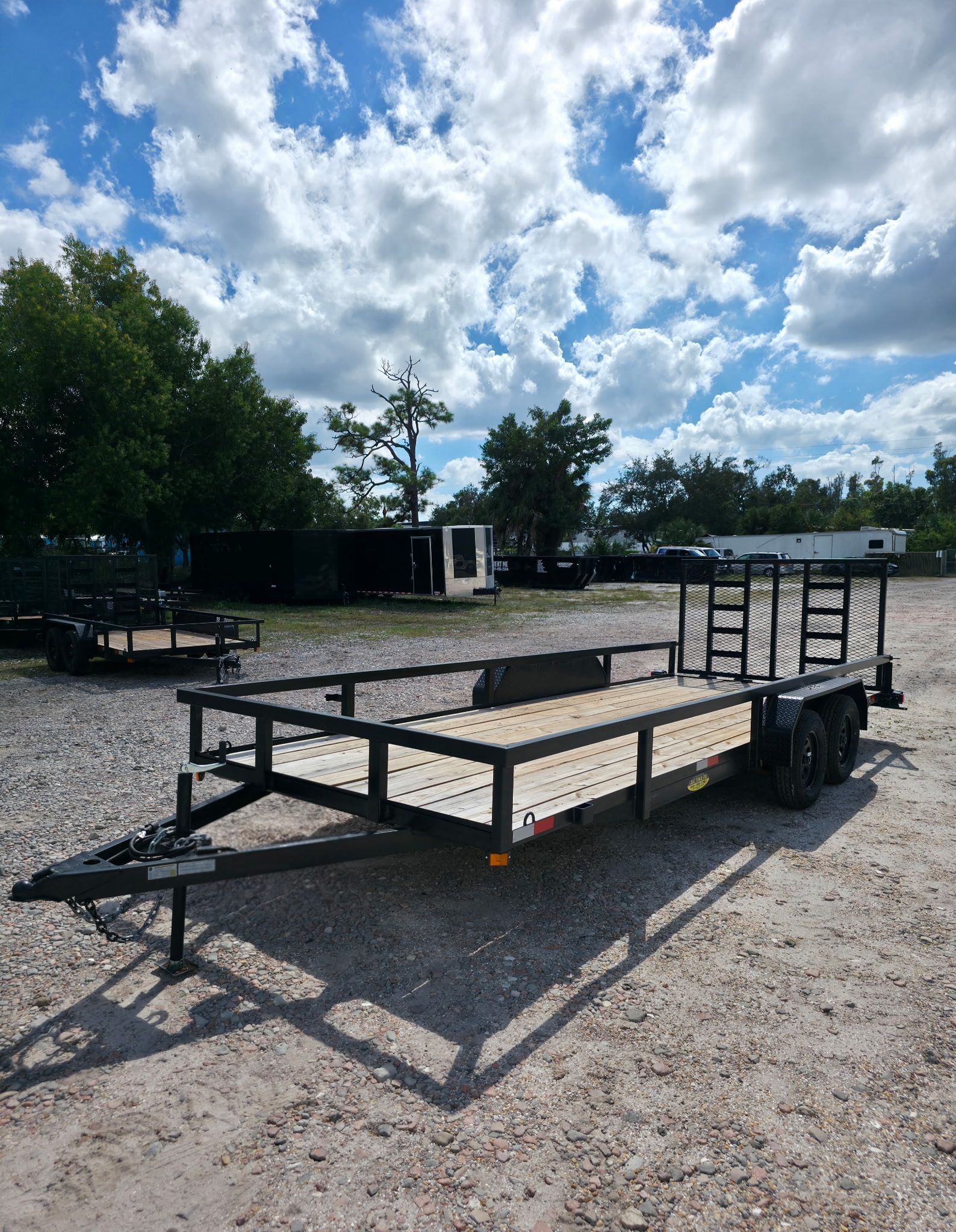 Black utility trailer on gravel with a sunny, cloudy sky in the background.