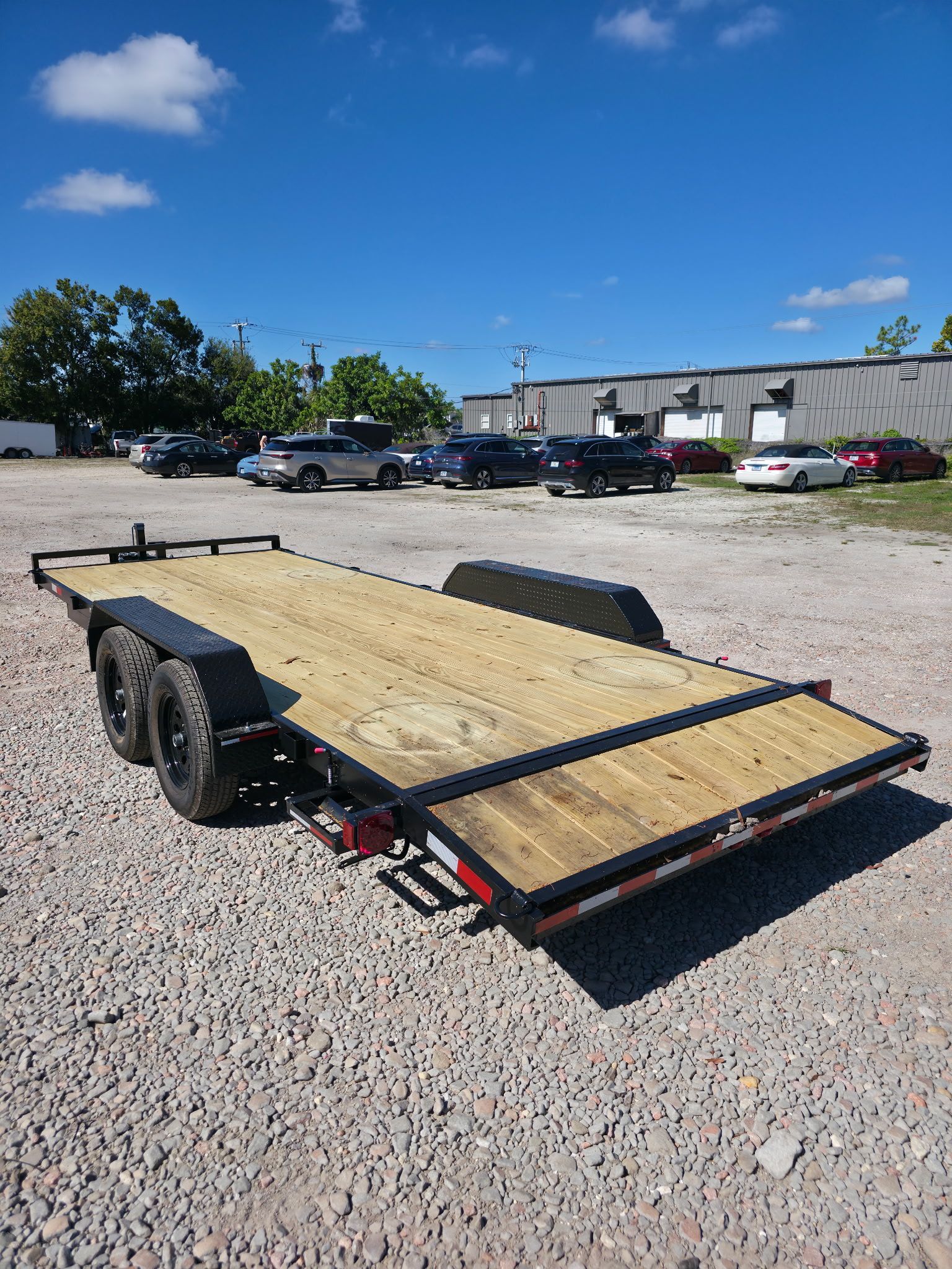 A black and wooden car trailer on a gravel lot under a blue sky, with cars parked in the background.