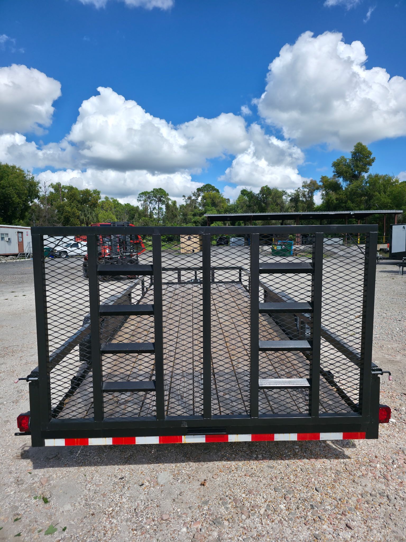 Black utility trailer with perforated metal sides and red/white reflective bumper, on gravel, blue sky.