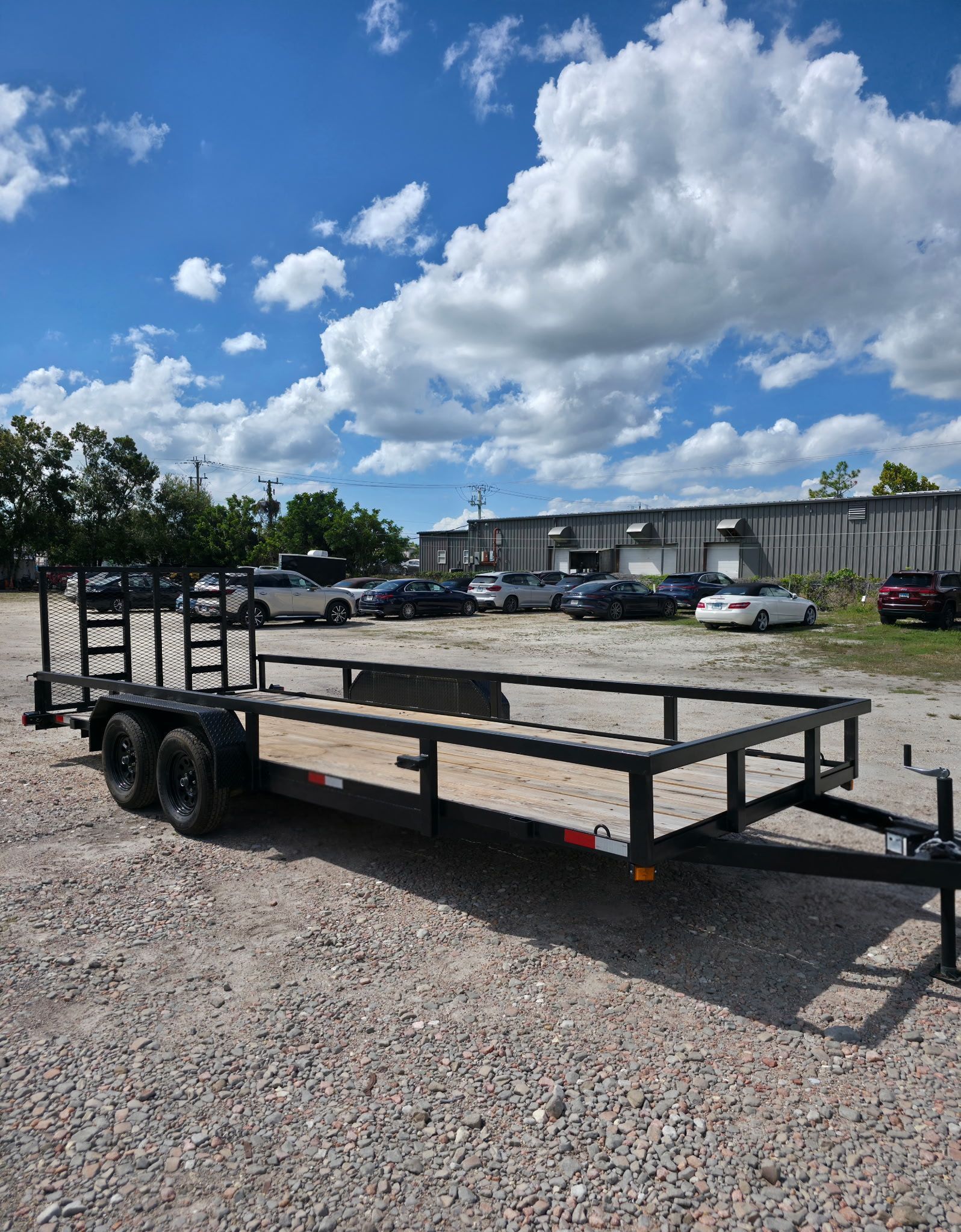 Black trailer parked on gravel, with cars and a building in the background under a blue sky with clouds.
