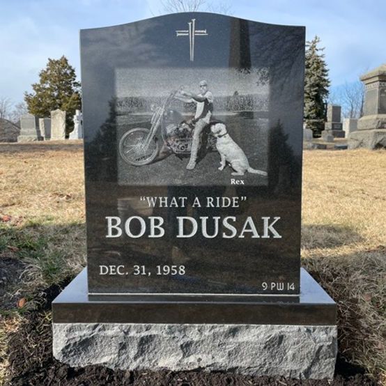 A black gravestone with a laser-etched image of a man on a motorcycle next to his dog. The inscription reads
