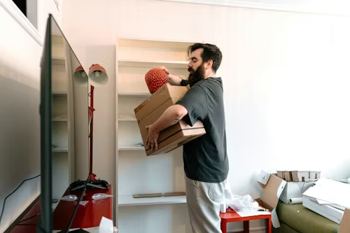 Man carrying stacked boxes through a doorway in a small room with a red scooter and scattered furniture