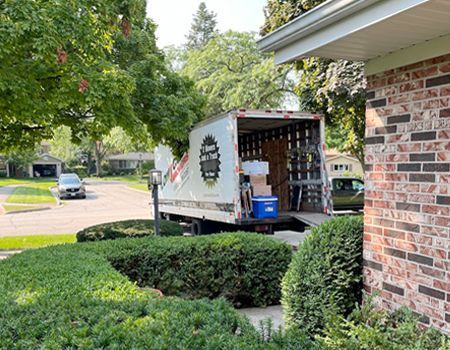 Moving truck parked in a driveway; boxes inside, next to a brick house with green bushes and trees.