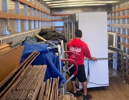 Man in red shirt pushing a refrigerator into a moving truck, full of boxes and furniture.